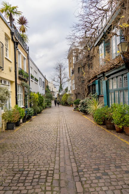 A narrow cobblestone pedestrian street lined with residential buildings on both sides, featuring a mix of modern and traditional architectural styles. On the left side, light-colored buildings with large windows, some with small balconies and decorative elements, are partially obscured by potted plants and greenery placed along the edge of the pavement. To the right, older buildings with darker brick facades and more extensive greenery, including climbing plants and trees, are visible; some parts of the facades are overgrown with vegetation. The street surface is composed of small, uneven cobblestones arranged in a linear pattern, with a central groove for drainage. In the background, a few people are seen walking away from the camera, and the sky overhead is cloudy with soft, diffused light, creating a calm, quiet atmosphere typical of an urban residential alley used for private access and rubbish collection, sometimes associated with independent waste disposal services like Rubbish Collection Brompton.