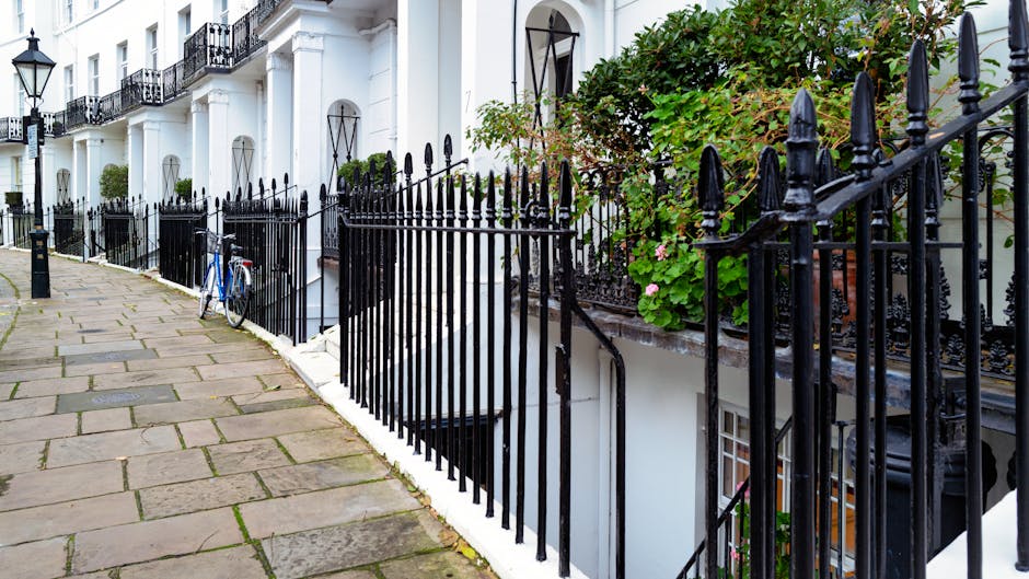A cobblestone courtyard enclosed by white terraced townhouses on both sides, with potted plants and small trees arranged along the front walls. The right side features a wooden fence with climbing greenery, while the left side has a black iron railing with additional potted plants on the ledge. Several benches made of wood and metal are positioned along the edges of the space, and small decorative bushes are placed in pots on the ground. The background shows an ornate white building with large windows and a decorative entrance, beneath a partly cloudy sky with some visible tree branches. The overall scene is well-lit, highlighting the textured cobblestones and lush foliage, reflecting a quiet residential setting possibly prepared for or involved in a private waste clearance process, such as the services offered by Rubbish Collection Brompton for alternative rubbish removal.