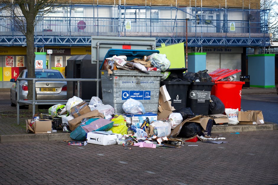 A collection of overflowing rubbish bins and scattered waste materials positioned on a paved sidewalk in front of a commercial building. The scene includes a large grey mixed paper and card bin with its lid open, revealing a jumble of cardboard, paper, and plastic waste spilling onto the ground. Surrounding this bin are multiple black and red waste bags, some tied and others torn open, with additional cardboard boxes and packaging materials strewn around the base. To the left, a silver car is parked behind a waist-high metal barrier that separates the rubbish area from the vehicle. The background features a retail storefront with closed shutters, signage, and a section of scaffolding along the upper part of the building, indicating ongoing construction or maintenance work. The lighting suggests overcast weather, contributing to a neutral, everyday urban atmosphere, aligning with the context of private waste collection or rubbish removal services offered by pathways such as Rubbish Collection Brompton in local shops or commercial settings.