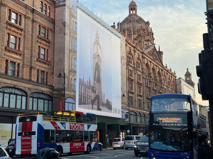 A busy urban street scene featuring historic multi-storey buildings with ornate stone facades and large windows, one of which displays a tall, vertical advertisement for Dior with a fashionable model holding a handbag, set against a partly cloudy sky. In the foreground, a double-decker bus in black and blue is heading to Kensington, with a digital sign on the front displaying route information, while a white and red tour bus labeled 'TOOT Bus' is parked nearby. Several cars are visible on the street, and pedestrians, some wearing face masks, walk along the sidewalk, illustrating a typical city environment with active public transportation and commercial advertising, all within a setting that reflects an active, vibrant London area with historic architecture and modern signage.