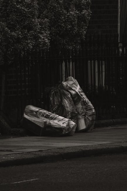 In the foreground, three large black rubbish bags made of thick plastic are placed on a paved sidewalk, leaning against a black metal fence with vertical bars. The bags appear to be filled with waste, with some areas showing crumpled and uneven surfaces, indicating they are bulging with contents. Behind the fence, dense foliage of leafy trees and shrubs is visible, adding texture and greenery to the background. The scene is dimly lit, suggesting overcast weather or low natural light, and the overall environment appears to be an urban or suburban street setting. The rubbish collection site may relate to private waste disposal or alternative rubbish removal services, as indicated by the presence of the bags in this outdoor area. The image’s composition emphasizes the contrast between the dark fencing, leaves, and the shiny, crumpled plastic of the rubbish bags, capturing a typical scene of waste awaiting collection outside a residential property or an informal rubbish disposal spot, relevant to rubbish removal and waste management contexts.