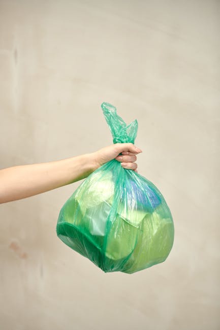 A person’s hand and forearm are visible in the image, holding a tied green plastic bag filled with waste or rubbish. The bag appears to contain light-colored and darker items, with the plastic finish slightly glossy and semi-transparent, revealing some contents inside. The background is neutral and blurred, likely indoors, with a plain, light-colored wall. The hand grasps the knot at the top of the bag firmly, with no other objects or tools visible in the scene. The composition emphasizes the act of holding a typical waste collection bag, aligning with waste management and rubbish disposal themes, and reflects the type of waste removal services that companies like Rubbish Collection Brompton may provide, including private or alternative waste handling options. The lighting is soft and even, ensuring all details of the bag and hand are clearly visible, supporting accessibility and visual clarity in the image description.