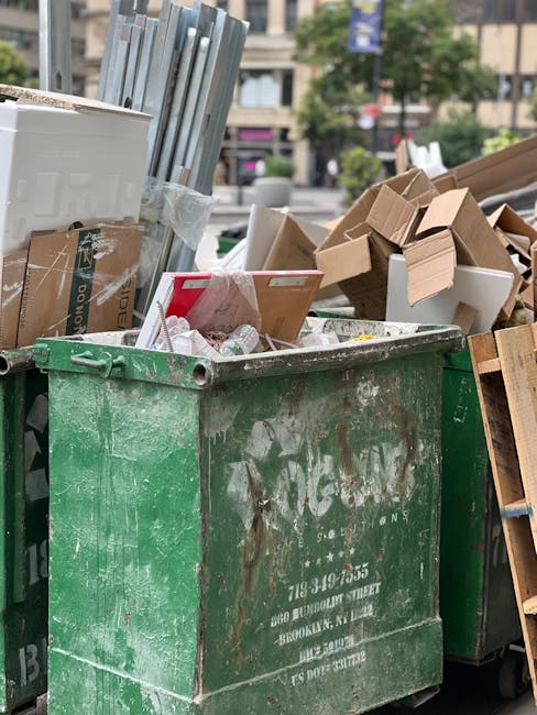 A large, weathered green industrial waste bin filled with a mixture of discarded cardboard boxes, some flattened and others still in their original shapes, along with crumpled paper and plastic packaging. The bin is situated outdoors on a paved urban area, with the surrounding environment including a busy street scene featuring trees, multi-storey buildings, and parked vehicles in the background. Several cardboard boxes are piled beside the bin, some with torn edges and open flaps, indicating recent or ongoing rubbish collection, possibly as part of private waste handling or alternative rubbish removal services. The bin shows signs of rust and dirt, highlighting its use for industrial or commercial waste collection. Natural daylight illuminates the scene, emphasizing the textures and colors of the cardboard, paper, and the worn surface of the bin, with an overall focus on facilitating effective rubbish disposal outside an apartment or commercial building, consistent with services provided by companies like Rubbish Collection Brompton for same-day rubbish removal in Brompton Road SW3.
