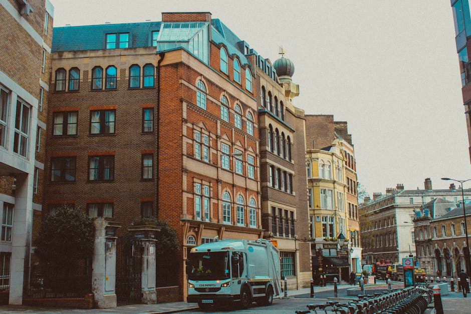 A street scene in an urban area featuring a row of mixed architectural style buildings, including a prominent red brick structure with decorative stonework and large arched windows. The buildings vary in height and design, with some having ornate facades and others with modern finishes. In the foreground, a white waste collection truck with a loading compartment is parked on the street, likely involved in private rubbish collection services. The street is lined with black bollards and a row of parked bicycles, and a few pedestrians are visible in the distance. The environment appears to be well-lit with natural daylight, and the overall scene reflects a typical city street where independent waste removal services, such as those offered by Rubbish Collection Brompton, operate to manage rubbish removal apart from local authority collection.