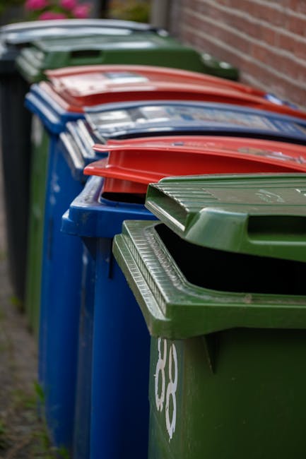 A large, weathered green industrial waste bin filled with a mixture of discarded cardboard boxes, some flattened and others still in their original shapes, along with crumpled paper and plastic packaging. The bin is situated outdoors on a paved urban area, with the surrounding environment including a busy street scene featuring trees, multi-storey buildings, and parked vehicles in the background. Several cardboard boxes are piled beside the bin, some with torn edges and open flaps, indicating recent or ongoing rubbish collection, possibly as part of private waste handling or alternative rubbish removal services. The bin shows signs of rust and dirt, highlighting its use for industrial or commercial waste collection. Natural daylight illuminates the scene, emphasizing the textures and colors of the cardboard, paper, and the worn surface of the bin, with an overall focus on facilitating effective rubbish disposal outside an apartment or commercial building, consistent with services provided by companies like Rubbish Collection Brompton for same-day rubbish removal in Brompton Road SW3.