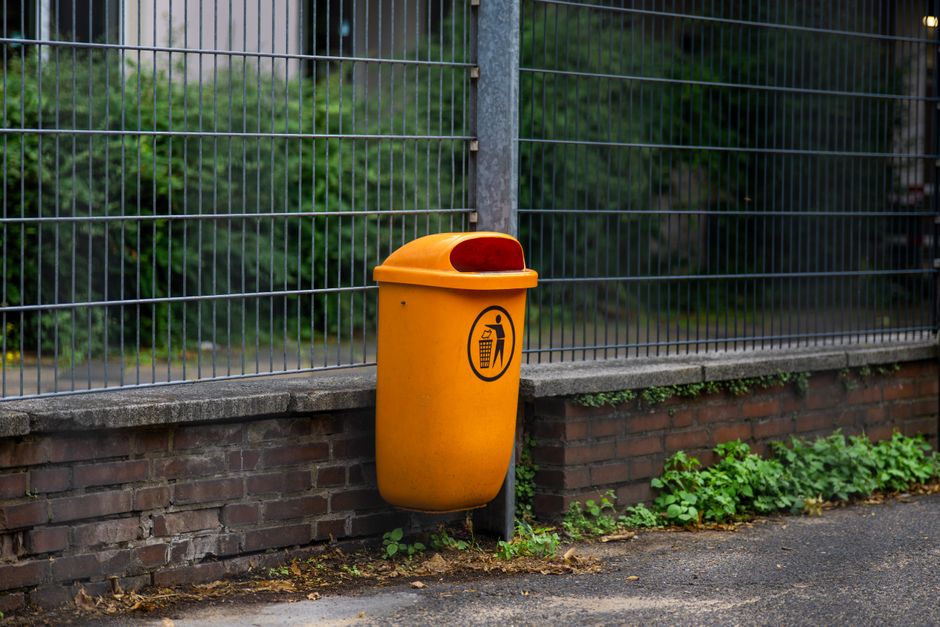 A bright orange outdoor waste bin is mounted on a brick wall section next to a metal fence with vertical and horizontal bars. The bin features a rounded top with a red lid and a black recycling symbol indicating general waste disposal. The surrounding environment includes a concrete pavement with some green plants and small weeds growing along the base of the wall. Behind the fence, there is lush greenery with trees and bushes, suggesting an outdoor setting possibly adjacent to a garden or park area. The scene is illuminated by natural daylight, creating a neutral, functional atmosphere typical of urban or suburban rubbish collection points. The placement of the bin near the fence indicates it may be part of a private waste collection or an alternative rubbish disposal setup, aligning with services such as those provided by Rubbish Collection Brompton for on-site waste management and disposal of garden or household waste.