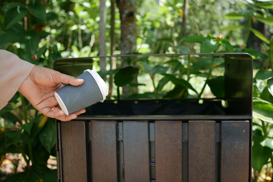 A bright orange outdoor waste bin is mounted on a brick wall section next to a metal fence with vertical and horizontal bars. The bin features a rounded top with a red lid and a black recycling symbol indicating general waste disposal. The surrounding environment includes a concrete pavement with some green plants and small weeds growing along the base of the wall. Behind the fence, there is lush greenery with trees and bushes, suggesting an outdoor setting possibly adjacent to a garden or park area. The scene is illuminated by natural daylight, creating a neutral, functional atmosphere typical of urban or suburban rubbish collection points. The placement of the bin near the fence indicates it may be part of a private waste collection or an alternative rubbish disposal setup, aligning with services such as those provided by Rubbish Collection Brompton for on-site waste management and disposal of garden or household waste.
